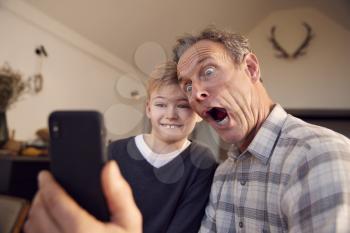Grandson With Grandfather Pulling Faces And Taking Selfie On Mobile Phone At Home