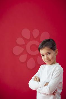 Portrait Of Young Boy With Folded Arms Against Red Studio Background Smiling At Camera