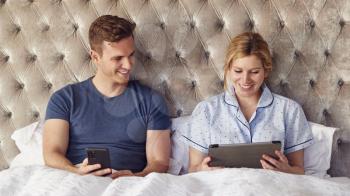 Couple At Home In Bed Self Isolating Using Digital Tablet And Mobile Phone During Covid 19 Lockdown