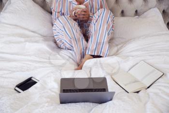 Close Up Of Woman Wearing Pyjamas Sitting On Bed With Laptop And Diary Working From Home