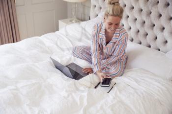 Businesswoman In Pyjamas Sitting On Bed With Laptop And Mobile Phone Working From Home In Lockdown