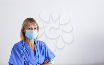 Studio Portrait Of Female Nurse Wearing Scrubs And PPE Face Mask And Gloves Against White Background