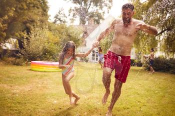 Father And Daughter Run Through Water From Garden Sprinkler Having Fun Wearing Swimming Costumes