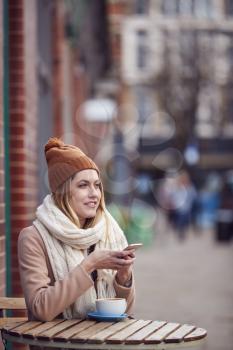 Female Customer Sending Text Message Sitting Outside Coffee Shop On Busy City High Street In Fall