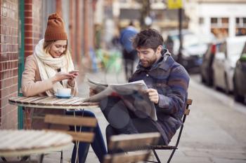 Couple On Date Sitting Outside Coffee Shop On High Street Using Mobile Phone And Reading Newspaper
