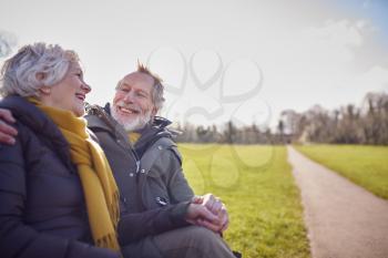 Loving Senior Couple Sitting On Seat Enjoying Autumn Or Winter Walk Through Park Together