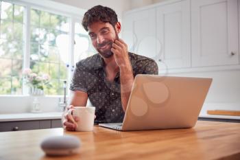 Man In Kitchen Using Digital Smart Speaker Whilst Working From Home With Laptop During Pandemic