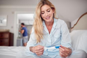 Smiling Woman Wearing Pyjamas In Bedroom Holding Positive Pregnancy Test