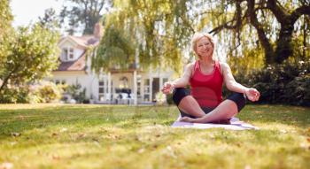 Portrait Of Senior Woman At Home In Garden Wearing Fitness Clothing Sitting On Yoga Mat