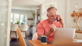 Retired Man On Phone At Home In Kitchen Using Laptop Celebrating Good News Or Winning