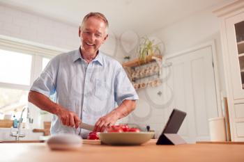 Retired Man Making Meal In Kitchen Asking Smart Speaker In Foreground Question