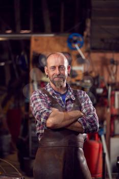 Portrait Of Mature Male Blacksmith Standing In Forge With Folded Arms