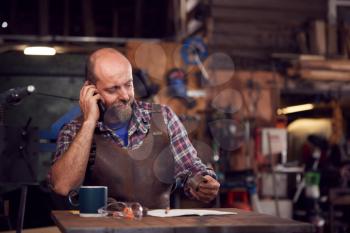 Mature Male Blacksmith Working On Design In Forge Whilst Using Mobile Phone