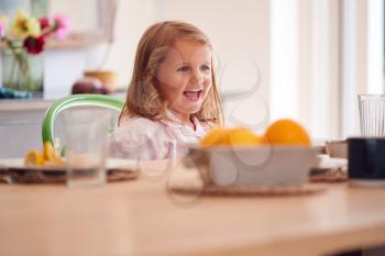 Smiling Young Girl Wearing Pyjamas Sitting At Table In Kitchen At Home Waiting For Breakfast