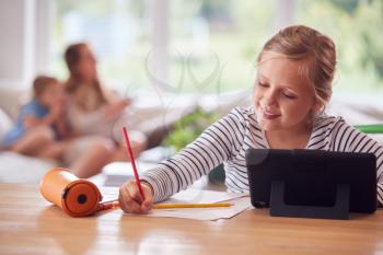 Girl Sitting At Table With Laptop Home Schooling During Health Pandemic With Family In Background
