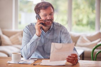 Smiling Man On Phone Call Sitting At Table At Home Reviewing Domestic Finances