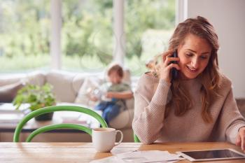 Mother On Phone Call At Home With Digital Tablet Reviewing Domestic Finances With Son In Background
