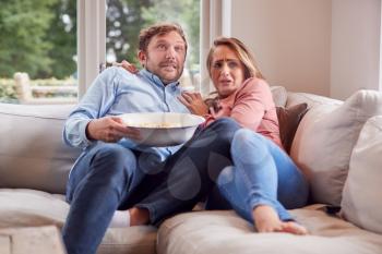 Couple On Date Night Sitting On Sofa At Home Watching Scary Movie On TV With Popcorn