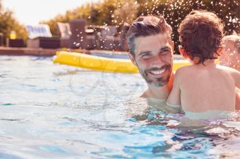 Family With Young Son Having Fun With Inflatable On Summer Vacation In Outdoor Swimming Pool