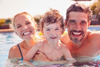 Portrait Of Family With Young Son Having Fun On Summer Vacation In Outdoor Swimming Pool