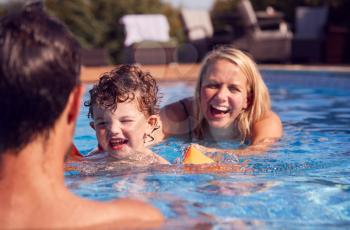 Family In Outdoor Pool On Summer Vacation Teaching Son To Swim With Inflatable Armbands