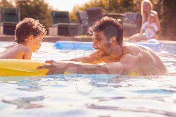 Father And Son Have Fun Playing In Outdoor Pool On Vacation As Mother And Baby Watch From Side