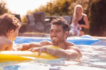 Father And Son Have Fun Playing In Outdoor Pool On Vacation As Mother And Baby Watch From Side