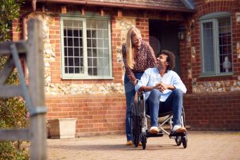 Mature Couple With Man Sitting In Wheelchair Being Pushed By Woman Outside Home