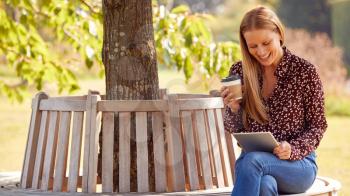 Mature Woman Sitting On Bench Under Tree In Summer Park Using Digital Tablet