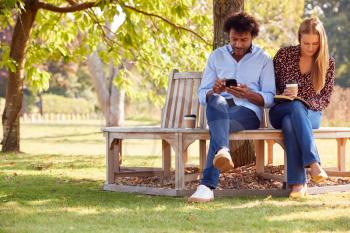 Mature Couple Relaxing Sitting On Park Bench Under Tree Reading Books And Checking Mobile Phone