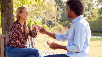 Mature Man Kneeling And Proposing To Surprised Woman Sitting In Park With Engagement Ring In Box