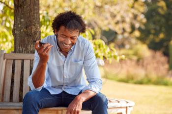 Mature Man Sitting On Park Bench Under Tree In Summer Talking On Mobile Phone Using Microphone
