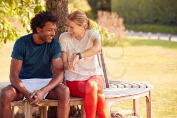 Couple Wearing Fitness Clothing Sitting On Seat Under Tree Checking Activity Monitor On Smartwatch