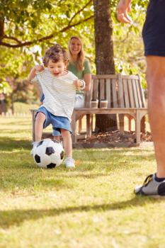 Family With Young Son Having Fun In Park Playing Football And Sitting On Seat Under Tree
