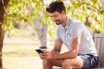 Man Wearing Summer Shorts Sitting On Park Bench Under Tree With Takeaway Coffee Using Mobile Phone