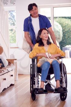 Asian Man Pushing Wife In Wheelchair At Home Back From Shopping Trip With Bag