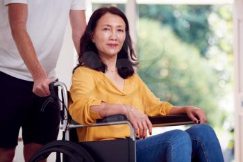 Portrait Of Asian Woman In Wheelchair Being Pushed By Carer At Home