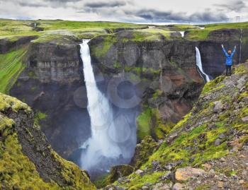 Spectacular waterfall Hayfoss in Iceland. Delighted woman - tourist in blue jacket standing on the edge of the cliff