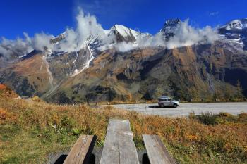 Snow-capped Alps in beautiful autumn day. Table and benches for picnic on the side of the  road. Great highway winds between hillsides yellowed