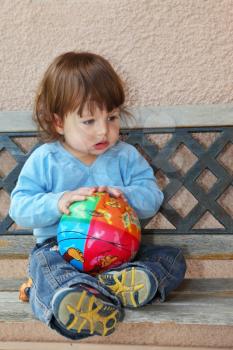 The charming little boy in dark blue jeans and blue sweater sits on to a garden bench with a bright multi-coloured ball in hands