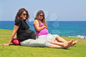 Two charming young pregnant women have a rest on a lawn at the sea.