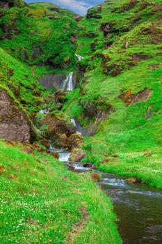  July in Iceland. Picturesque cascade multi-stage waterfall. Mountains covered with green grass and moss