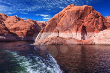 Red sandstone hills surround the lake. Foamy trace of a motor boat crosses the emerald waters. Lake Powell on the Colorado River