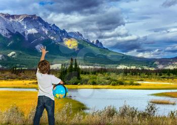 Magnificent landscape in the Rocky Mountains. Handsome boy with a globe in his hands is delighted with the mountains, clouds and lakes. The concept of ecological and active tourism