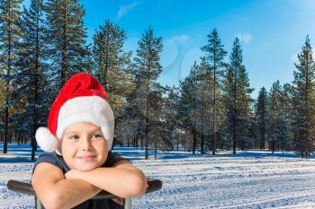 Charming boy in a red Santa Claus hat is smiling in a snow-covered winter forest. Waiting for Christmas