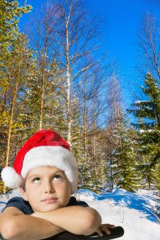 Charming boy in a red Santa Claus hat is smiling in a snow-covered winter forest. Cold winter sunset. Waiting for the New Year