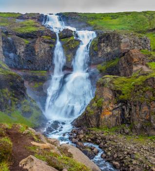  Cold and rainy July in Iceland. Powerful streams of cascade falls with a roar break against huge stones. Concept of active and extreme tourism