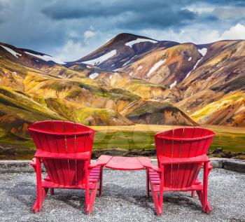 Two convenient red chaise lounges connected by small table stand for tourists.  Travel to Iceland in the July. Multi-colored mountains from mineral rhyolite are lit with sun