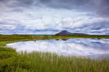 In the smooth water of cold lake reflects cloudy sky. Summer Iceland. Small lake surrounded by green meadow