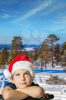 Cold winter evening. In a snowy forest, adorable boy in a red Santa Claus hat is waiting for Christmas
